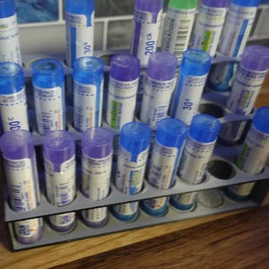 May include: A gray plastic storage rack holding multiple cylindrical tubes with blue and purple caps. Each tube features a white label with printed text, including numbers like 30c and 200ck, and barcodes. The rack is organized in rows.