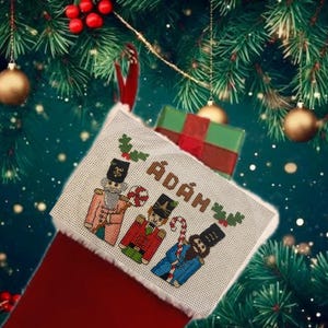 Puede incluir: Un calcetín navideño rojo con puño blanco, bordado con tres figuras de cascanueces y el nombre "ÁDÁM". Un pequeño regalo envuelto es visible dentro del calcetín, con un fondo de ramas y adornos de árbol de Navidad.