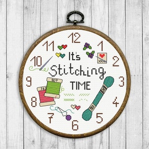 Könnte beinhalten: Ein weißer Stickrahmen mit einem braunen Holzrahmen. Der Rahmen zeigt ein Zifferblatt mit den Zahlen 1 bis 12 und dem Text "It's Stitching Time". Das Zifferblatt ist mit verschiedenen Stickartikeln verziert, darunter Nadeln, Garnrollen und Scheren.