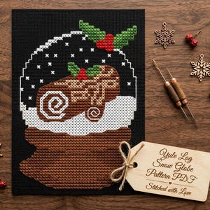 May include: A cross-stitch pattern of a snow globe with a Yule log inside, featuring green holly and red berries. The design is on a black background. A wooden tag reads "Yule Log Snow Globe Pattern PDF Stitched with Love."