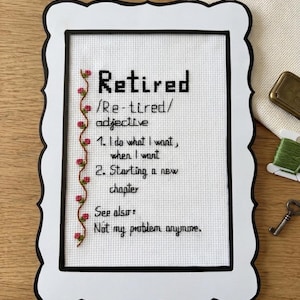 May include: A cross-stitch embroidery piece in a white scalloped frame with black trim. The white fabric features black text reading: Retired /Re-tired/ adjective 1. I do what I want, when I want 2. Starting a new chapter See also: Not my problem anymore.