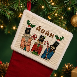 May include: Red Christmas stocking with a cream embroidered panel featuring three nutcracker figures and the name "ÁDÁM" in cross-stitch. The stocking has a white faux fur trim, with Christmas ornaments and greenery in the background.