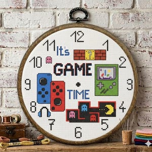Può includere: Un orologio rotondo a punto croce con cornice in legno. Il quadrante presenta le parole "It's GAME TIME" e grafica di videogiochi pixelati. I numeri sono neri e l'orologio è appeso a un muro di mattoni bianchi.