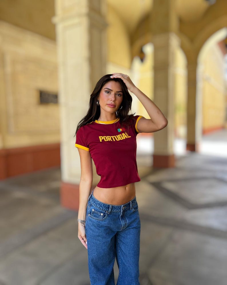 May include: A woman wearing a maroon crop top with yellow trim and the word "PORTUGAL" printed on the front. The top has a small Portuguese flag embroidered on the chest. She is also wearing blue jeans.