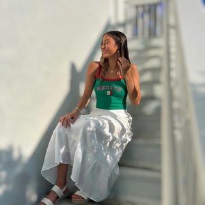 May include: A woman wearing a green tank top with the word "MEXICO" and the Mexican flag printed on it. She is wearing a white skirt and white sandals. She is sitting on a set of white stairs.