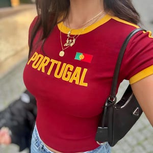 May include: Red t-shirt with yellow trim and the word "PORTUGAL" in yellow letters. The shirt features a small embroidered Portuguese flag. The person is wearing a gold necklace with a pendant.