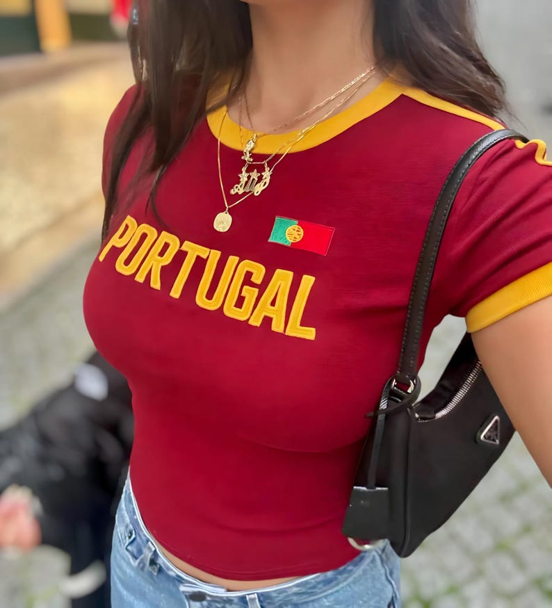 May include: Red t-shirt with yellow trim and the word "PORTUGAL" in yellow letters. The shirt features a small embroidered Portuguese flag. The person is wearing a gold necklace with a pendant.