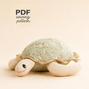 May include: A stuffed turtle toy featuring a sage green shell with a white floral pattern and a tan fabric body. The turtle has a stitched face with black eyes and a red mouth. Text above the turtle reads PDF sewing pattern.