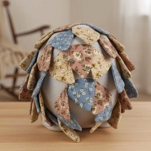 May include: A decorative, handmade fabric object with a rounded shape. The object is covered in petal-shaped fabric pieces in shades of blue, pink, and beige, each with a floral pattern. The base is a light cream color.
