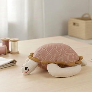 Könnte beinhalten: Ein Stofftier-Schildkröte in Rosa und Creme. Die Schildkröte hat einen strukturierten Panzer, cremefarbene Flossen und ein lächelndes Gesicht. Nähutensilien, darunter Garnrollen und ein Stoffquadrat, liegen auf dem Tisch.