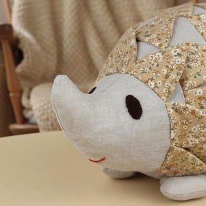 May include: A handmade hedgehog plush toy with a light gray face, brown eyes, and a small red smile. The body is covered in a floral patterned fabric with shades of tan, cream, and green. The toy is resting on a light tan surface.