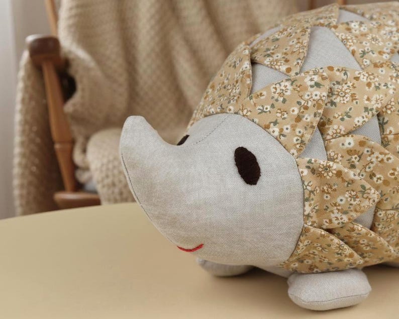 May include: A handmade hedgehog plush toy with a light gray face, brown eyes, and a small red smile. The body is covered in a floral patterned fabric with shades of tan, cream, and green. The toy is resting on a light tan surface.