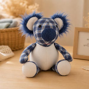Könnte beinhalten: Ein Koala-Stofftier mit blau-weißem Karomuster an Kopf, Armen und Beinen. Es hat einen einfarbig weißen Bauch, weiße Innenohren und eine dunkelblaue, texturierte Nase. Die Ohren sind mit blauem Kunstfell akzentuiert und es hat schwarze Augen.