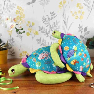 May include: Two plush sea turtle toys, one green and one blue, with colourful ocean-themed patterns. The green turtle is larger and has a pink, yellow, and purple shell. The blue turtle is smaller and has a pink, yellow, and purple shell. Both turtles have smiling faces and are made of soft fabric.
