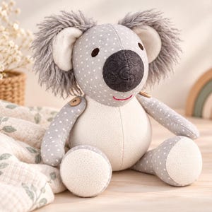 May include: A plush koala toy featuring a gray body with white polka dots, a cream-colored belly, and dark gray ears with shaggy faux fur. The koala has a dark gray nose with a swirl pattern, small black eyes, and a stitched red mouth.