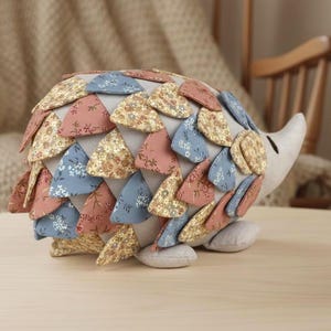 May include: A stuffed hedgehog toy with a cream-colored body and a snout. The back is covered in triangular fabric pieces in shades of pink, blue, and yellow with floral patterns. The toy is sitting on a wooden surface.
