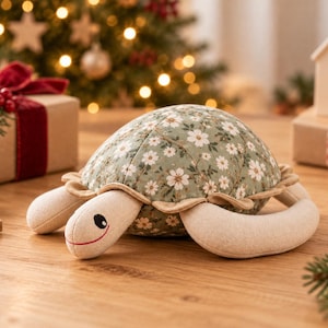 Könnte beinhalten: Ein Plüsch-Schildkrötenspielzeug mit einem gesteppten, salbeigrünen Panzer mit weißem Blumenmuster. Kopf und Flossen bestehen aus hellbeigem Stoff. Die Schildkröte hat ein gesticktes schwarzes Auge und einen dünnen roten Mund.
