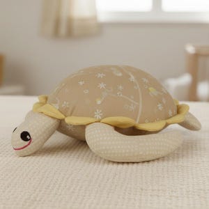 Puede incluir: Un peluche de tortuga beige con caparazón estampado y ribetes amarillos. La tortuga tiene una cara sonriente con ojos negros y boca cosida en rojo. Juguete para niños.