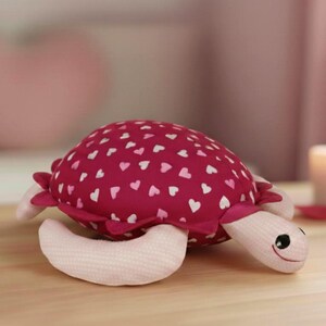 Può includere: Un giocattolo a forma di tartaruga di peluche con un guscio bordeaux decorato con cuori bianchi. La tartaruga ha arti rosa chiaro e un viso sorridente. Il giocattolo è su una superficie di legno, con una candela accesa sullo sfondo.