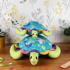 May include: Two plush sea turtle toys stacked on top of each other. The bottom turtle is green with a blue, yellow, and pink shell with a sea life pattern. The top turtle is green with a blue shell with stars. Both turtles have purple eyes and a pink nose.