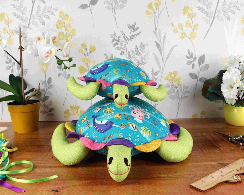 May include: Two plush sea turtle toys stacked on top of each other. The bottom turtle is green with a blue, yellow, and pink shell with a sea life pattern. The top turtle is green with a blue shell with stars. Both turtles have purple eyes and a pink nose.