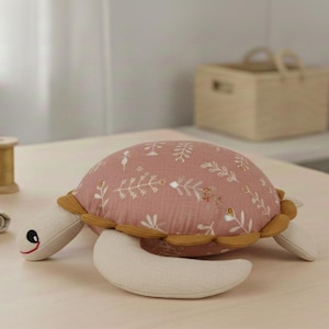 Può includere: Un peluche a forma di tartaruga con un guscio rosa con un motivo floreale bianco. La tartaruga ha pinne e testa color crema, con un bordo giallo attorno al guscio. Gli occhi sono neri con un sorriso rosso.