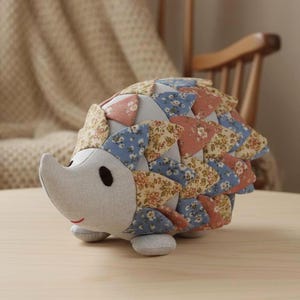 Op de afbeelding: Een handgemaakt knuffel-egel. De egel heeft een lichtgrijs gezicht en buik, met een patchwork rug in tinten blauw, roze en beige bloempatronen. Het heeft kleine zwarte ogen en een gestikte rode glimlach. Het speelgoed staat op een houten oppervlak.