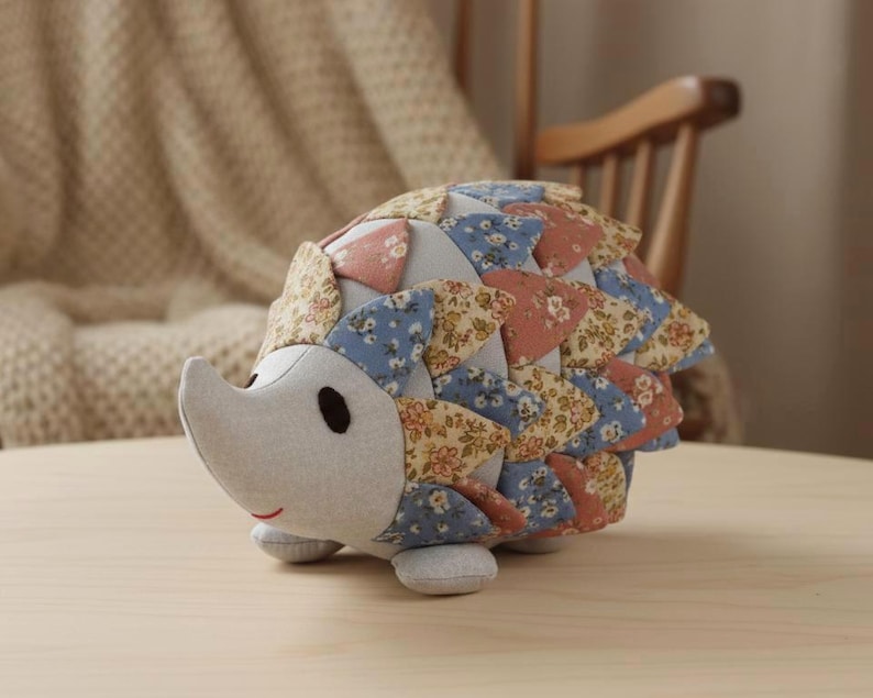 May include: A handmade stuffed hedgehog toy. The hedgehog has a light gray face and belly, with a patchwork back in shades of blue, pink, and beige floral patterns. It has small black eyes and a stitched red smile. The toy is sitting on a wooden surface.