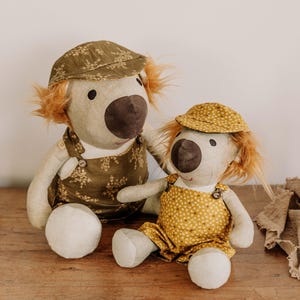 Puede incluir: Dos muñecos de león de peluche con melenas naranjas y narices marrones. El muñeco más grande lleva una gorra y un peto con estampado floral verde. El muñeco más pequeño lleva una gorra y un peto de lunares amarillos. Ambos muñecos están sentados sobre una superficie de madera.