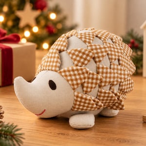Peut inclure: Une peluche en forme de hérisson avec un corps en tissu crème, un museau pointu, des yeux brodés marron foncé et une petite bouche cousue en rouge. Le dos est recouvert de pièces de tissu triangulaires superposées à motif vichy marron et blanc.