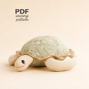 May include: A stuffed turtle toy featuring a sage green shell with a white floral pattern, a tan ruffled trim, and light beige limbs. The head has embroidered black eyes and a thin red mouth. Text in the upper left corner reads PDF sewing pattern.