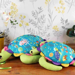 May include: Two plush sea turtle toys with colourful ocean-themed fabric. The larger turtle is lying on its back with its legs in the air. The smaller turtle is lying on its side with its head resting on the larger turtle's shell.