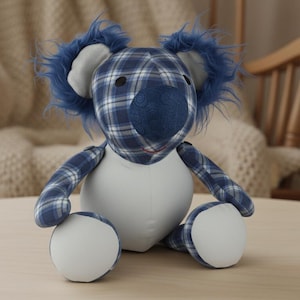 May include: A plush koala bear with a blue plaid pattern on its head, arms, and legs. The koala has a white body and white circular feet. The ears are trimmed with blue fur. The koala is sitting on a light brown surface.