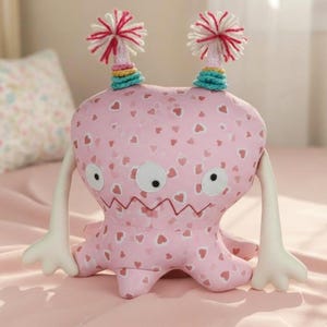 Puede incluir: Un juguete de monstruo de peluche rosa con patrones de corazón, brazos blancos y ojos grandes blancos. El monstruo tiene dos antenas de pompón con hilo rojo y blanco. El juguete está sobre una superficie rosa.