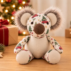 Peut inclure: Une peluche koala en tissu crème ornée d'un motif festif représentant des camions rouges transportant des sapins de Noël, des pins verts et des étoiles dorées. Le koala possède de grandes oreilles grises duveteuses, un nez brun et des yeux noirs.