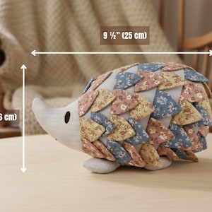 May include: A handmade fabric hedgehog plush toy. The hedgehog is 9 1/2 inches (25 cm) long and 6 inches (16 cm) tall. It features a light gray face and belly, with a back covered in floral patterned fabric in shades of pink, blue, and yellow.