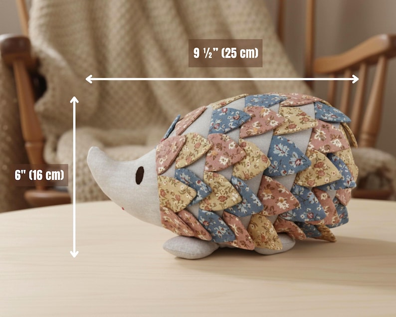 May include: A handmade fabric hedgehog plush toy. The hedgehog is 9 1/2 inches (25 cm) long and 6 inches (16 cm) tall. It features a light gray face and belly, with a back covered in floral patterned fabric in shades of pink, blue, and yellow.
