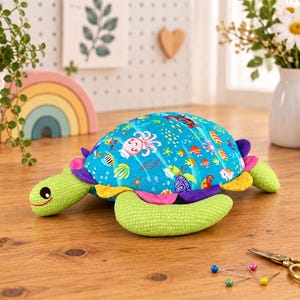 May include: A stuffed turtle pincushion featuring a bright blue shell patterned with colorful sea creatures including an octopus, crab, and fish. The turtle has a light green head and flippers with a white dot pattern and a ruffled edge.