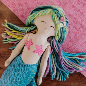 Pode incluir: Boneca de sereia feita à mão com um rosto sorridente, aplicações em forma de estrela e cabelo comprido de fio colorido. A cauda da sereia tem um padrão geométrico azul. A boneca é um brinquedo macio e de pelúcia.