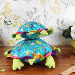 May include: Two stacked plush turtle toys with a blue background and colourful sea creatures. The top turtle has a purple, pink, and yellow shell. The bottom turtle has a purple, pink, yellow, and green shell.