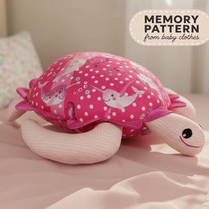 Peut inclure: Une peluche en forme de tortue avec une carapace rose vif ornée de pois blancs et d'illustrations de narvals et d'hippocampes. La tête et les nageoires sont en tissu rose clair avec une texture à pois blancs. Le texte indique MEMORY PATTERN from baby clothes.