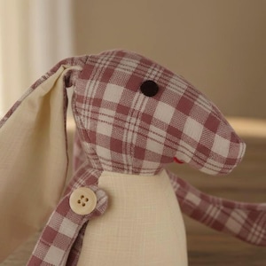 Puede incluir: Un conejo de peluche hecho a mano con un estampado de cuadros rosas y blancos. El conejo tiene orejas largas, un detalle de botón y un cuerpo color crema. El ojo del conejo es un pequeño botón marrón oscuro.