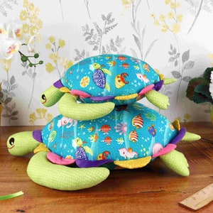May include: Two plush sea turtle toys stacked on top of each other. The bottom turtle is green with a blue and white shell with colourful fish and sea life designs. The top turtle is green with a blue shell with colourful fish and sea life designs.