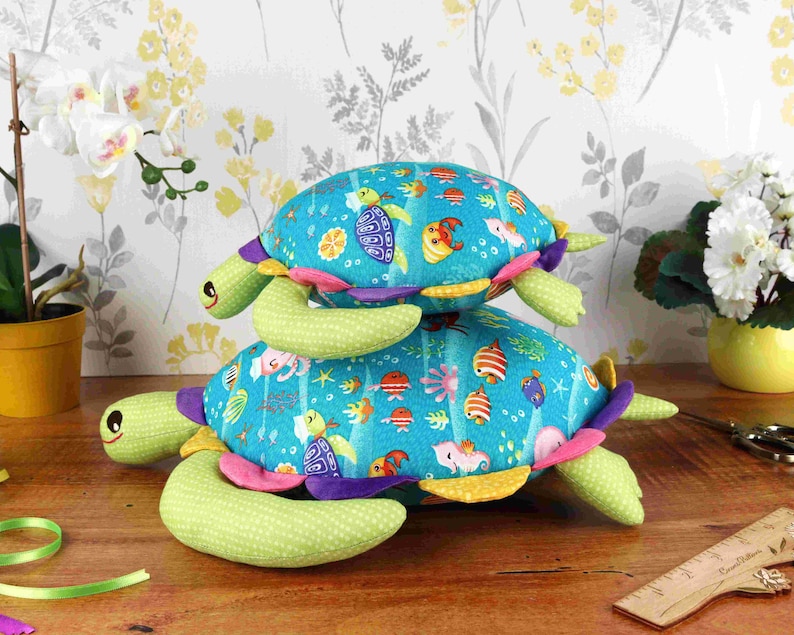 May include: Two plush sea turtle toys stacked on top of each other. The bottom turtle is green with a blue and white shell with colourful fish and sea life designs. The top turtle is green with a blue shell with colourful fish and sea life designs.