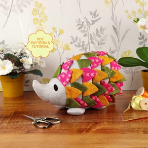 Puede incluir: Un patrón PDF y un tutorial para coser un juguete de peluche de erizo. El erizo está hecho de tela en tonos de verde, rosa y amarillo. El juguete está sentado en una mesa de madera con un par de tijeras, cinta y plantas en macetas.