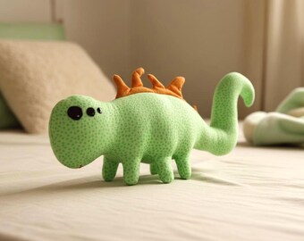 May include: A light green stuffed dinosaur toy with orange spikes on its back. The dinosaur has a long, curved tail and small legs. The toy is made of a soft, dotted fabric and has large black eyes.