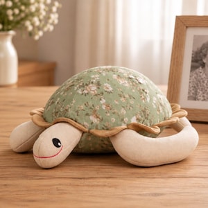 May include: A plush turtle toy featuring a sage green shell with a white and pink floral pattern. The turtle has a cream-colored head and limbs, with a stitched black eye and a thin red line for a mouth. A tan fabric ruffle borders the shell.