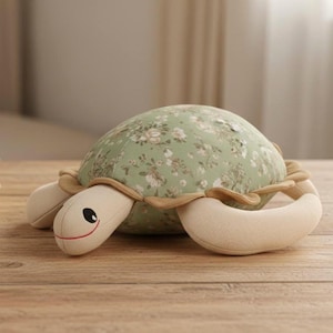 Pode incluir: Um brinquedo de pelúcia de tartaruga com uma carapaça verde com estampa floral e membros bege. A tartaruga tem um rosto sorridente com olhos pretos e uma boca costurada em vermelho. O brinquedo está em uma superfície de madeira.