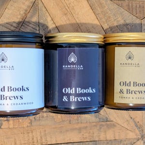 May include: Three glass jar candles with black, gold, and silver lids. Each jar has a label with the text "Old Books & Brews" and the brand name "Kandella & Co." The labels are white, navy blue, and tan.