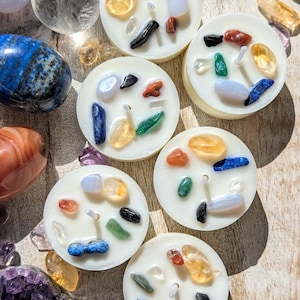 May include: A collection of white, circular candles embedded with colorful gemstones. Various polished stones, including blue lapis lazuli and amethyst, surround the candles. The gemstones are in shades of blue, green, yellow, red, and black.